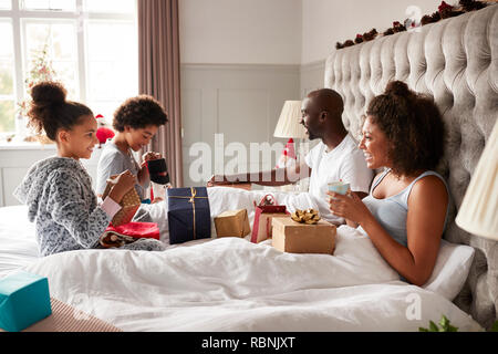 Junge Kinder öffnen Geschenke auf dem Bett der Eltern am Weihnachtsmorgen, während ihre Eltern im Bett sitzen beobachten, Seitenansicht
