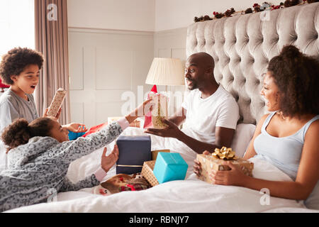 Jungen gemischten Rennen Familie sitzt auf dem Bett der Eltern geben jedem anderen Geschenken auf Weihnachten Morgen, in der Nähe Stockfoto
