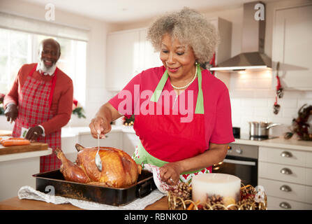 Ältere schwarze Frau basting ein Braten in der Türkei in Vorbereitung für das Weihnachtsessen, ihr Ehemann Zerkleinern von Gemüse im Hintergrund, vorne, Stockfoto