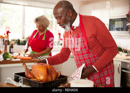 Älteren schwarzen Mann basting Truthahn in Vorbereitung für das Weihnachtsessen, seine Frau Zerkleinern von Gemüse im Hintergrund, in der Nähe Stockfoto