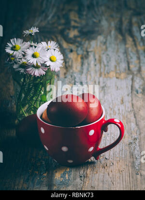 Ostern noch Leben auf dunklem Hintergrund. Hand gemalte rote Ostereier in retro Kanne und daisy flower bouquet auf rustikalen Holztisch Kunst Traditionelle Stockfoto