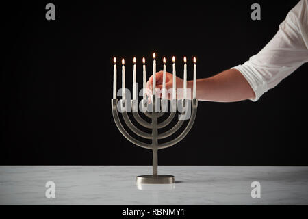 Hand Kerzenlicht auf Metall Hanukkah Menorah auf Marmor Oberfläche gegen Schwarz Studio Hintergrund Stockfoto