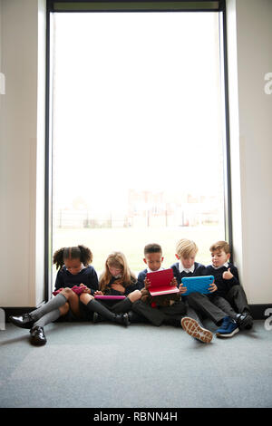 Grundschule Kinder gemeinsam sitzen auf dem Boden vor einem Fenster in einer Schule Korridor mit Tablet Pcs, vertikal Stockfoto