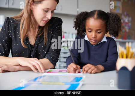 Junge weibliche Grundschullehrerin arbeitet auf eine mit einem Schulmädchen gerade Ihre an einem Tisch in einem Klassenzimmer schreiben, in der Nähe Stockfoto