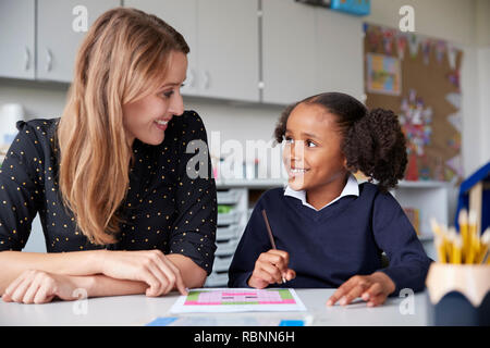 Junge weibliche Grundschullehrerin arbeitet auf einer mit einer Schülerin an einem Tisch in einem Klassenzimmer, beide schauen einander an, Lächeln, Nahaufnahme Stockfoto