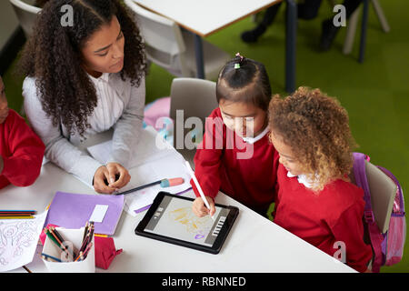 Ansicht von zwei Mädchen mit einem Tablet Computer am Tisch sitzen in einem Kleinkind Schule Klasse a mit Lehrerin ihnen helfen Stockfoto