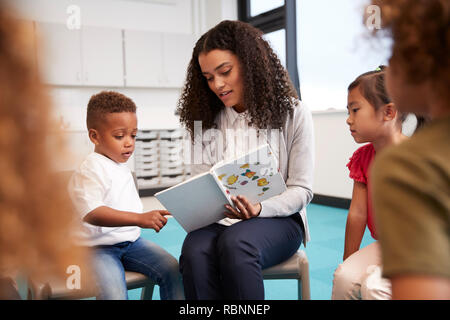 Infant School Junge in einem Buch von der Lehrerin gehalten, mit Kinder sitzen im Kreis auf Stühlen im Klassenzimmer, in der Nähe Stockfoto