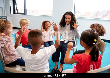 Eine Klasse für Kleinkinder Schulkinder sitzen auf Stühlen im Kreis im Klassenzimmer, die Hände und Lernen mit ihrer Lehrerin zu zählen, in der Nähe Stockfoto