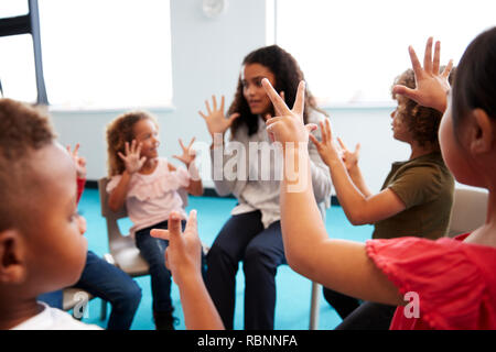 Eine Klasse für Kleinkinder Schulkinder sitzen auf Stühlen im Kreis im Klassenzimmer, die Hände und Lernen mit ihrer Lehrerin zu zählen, in der Nähe Stockfoto