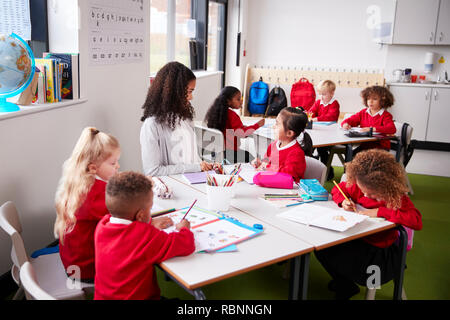 Junge weibliche Infant School Lehrer an einem Tisch sitzen in einem Klassenzimmer mit ihren Schülern Stockfoto