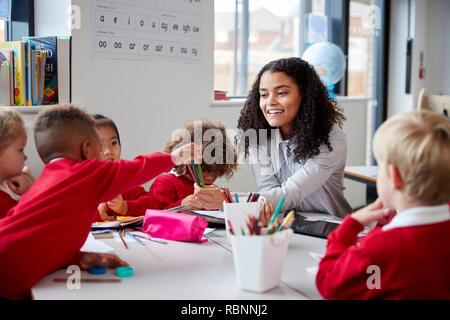 Vorderansicht des lächelnd weibliche Infant School Lehrer an einem Tisch im Klassenzimmer sitzen mit einer Gruppe von Schulkindern Stockfoto