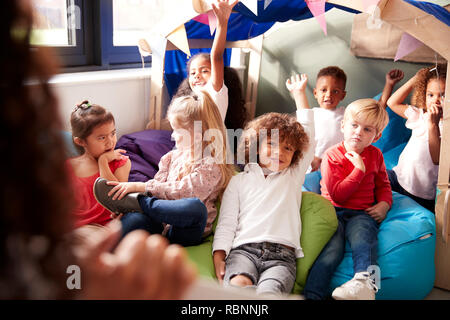 Blick über die Schulter des Kindes Schule Lehrer zeigen ein Buch zu einer Gruppe von Kindern sitzen auf Bohne Beutel in einer gemütlichen Ecke des Klassenzimmers, heben die Hände eine Frage, selektiven Fokus auf Antwort, in der Nähe Stockfoto