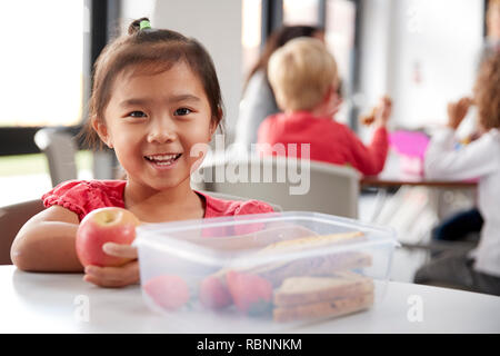 Junge chinesische Schülerin an einem Tisch sitzen während der Mittagspause im Kindergarten, mit einem Apfel und lächelnd, Nahaufnahme Stockfoto