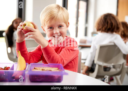 Junge weiße Schüler sitzen an einem Tisch und lächelnd einen Apfel in einem Kindergarten Klassenzimmer während seiner Mittagspause, in der Nähe Stockfoto