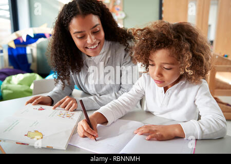 Weibliche Infant School Lehrer arbeitet auf einer mit einer jungen Schülerin an einer Tabelle schreiben in einem Klassenzimmer sitzen, vorne, Stockfoto