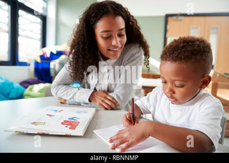 Weibliche Infant School Lehrer eine Arbeit an einem mit einem jungen Schüler, an einem Tisch in einem Klassenzimmer sitzen, vorne, Stockfoto