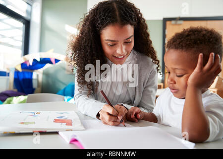 Weibliche Infant School Lehrer eine Arbeit an einem mit einem jungen Schüler, sitzen an einem Tisch mit ihm zu schreiben, in der Nähe Stockfoto