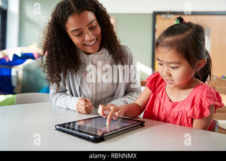 Weibliche Infant School Lehrer arbeitet auf einer mit einem chinesischen Schulmädchen, sitzen an einem Tisch in einem Klassenzimmer über einen Tablet-PC, in der Nähe Stockfoto