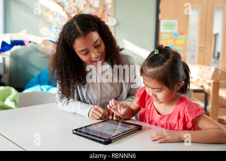 Weibliche Infant School Lehrer arbeitet auf einer mit einem chinesischen Schulmädchen, sitzen an einem Tisch in einem Klassenzimmer über einen Tablet-PC, in der Nähe Stockfoto