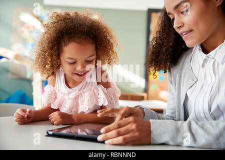 Weibliche Infant School Lehrer mit einem Tablet Computer arbeitet auf einer in einem Klassenzimmer mit einem Jungen gemischten Rennen Schulmädchen, selektiver Fokus, Nahaufnahme Stockfoto