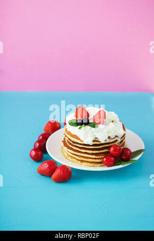 Pfannkuchen in einer Platte mit Sahne, Erdbeeren, Minze und Süßkirschen auf einem hellen Blau und Rosa Hintergrund. Kopieren Platz Vertikal Stockfoto