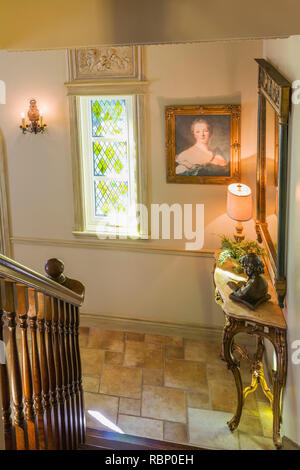 Eine Treppe aus braunem Eichenholz und eine antike Konsole mit Statue, eine beleuchtete Lampe im Eingangsbereich mit Buntglasfenster im Haus der Renaissance. Stockfoto