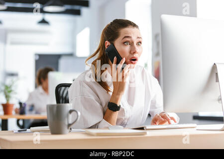 Enttäuscht, schockiert junge geschäftsfrau im Hemd an Ihrem Arbeitsplatz im Büro sitzen, gekleidet, Gespräch am Handy Stockfoto