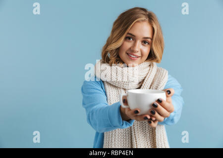 Bild der liebenswerten Frau 20 s eingewickelt in Schal und lächelnd Tasse mit Tee oder Kaffee über blauen Hintergrund isoliert Stockfoto