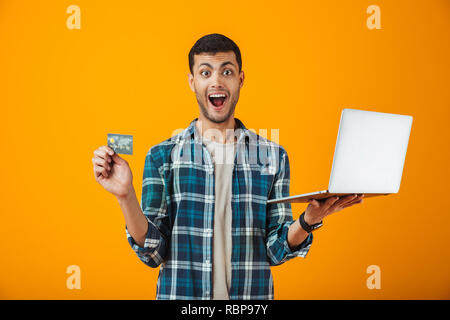 Fröhlicher junger Mann, der Plaid Shirt stehen über orange Hintergrund isoliert, mit Laptop Computer, Kunststoff Kreditkarte Stockfoto