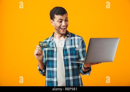 Fröhlicher junger Mann, der Plaid Shirt stehen über orange Hintergrund isoliert, mit Laptop Computer, Kunststoff Kreditkarte Stockfoto