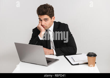 Müde, gelangweilt Young Business Mann, Klage über grauer Hintergrund isoliert, sitzen am Schreibtisch arbeiten am Laptop Stockfoto