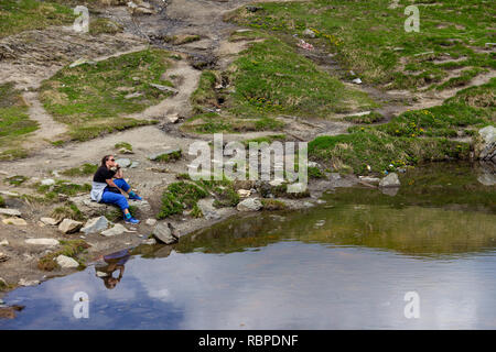 Eine Frau eine Sonnenbrille tragen und Wandern Kleidung sitzt nachdenklich am Seeufer Stockfoto