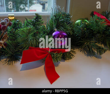 Weihnachtsgirlande mit einem roten Bogen und violetten und grünen Bommeln sitzt auf einem Felsvorsprung unter einem Fenster Stockfoto