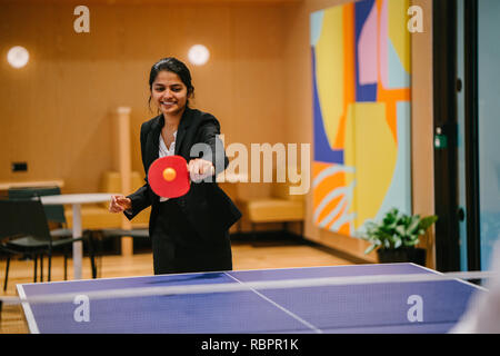 Eine junge indische asiatische Frau in einem Anzug lächelt und lacht, während Sie ping pong in Ihrer startup Office spielt. Sie hat viel Spaß mit einem Kollegen. Stockfoto