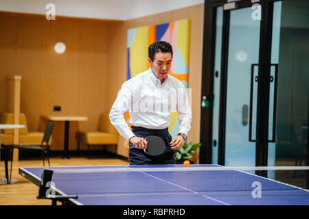 Eine asiatische Geschäftsmann ist Tischtennis spielen mit einem Kollegen in seinem Büro während des Tages. Er lächelte, als er das Spiel genießt. Stockfoto
