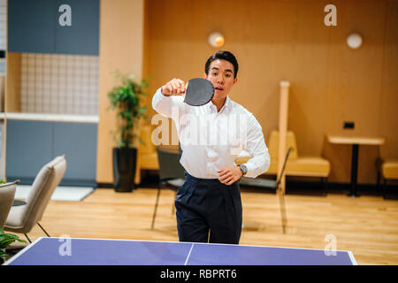 Eine asiatische Geschäftsmann ist Tischtennis spielen mit einem Kollegen in seinem Büro während des Tages. Er lächelte, als er das Spiel genießt. Stockfoto
