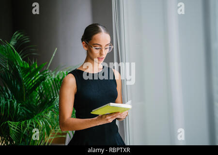 Portrait einer Jungen, Schönen und Eleganten blonden Frau Brille lesen ein Buch durch ein Fenster während des Tages. Sie ist professionell gekleidet. Stockfoto