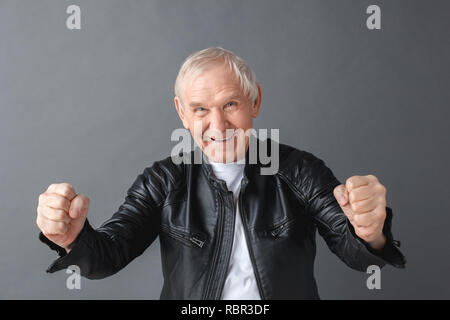Ältere Menschen tragen schwarze Lederjacke stehen isoliert auf graue Wand ballte die Fäuste suchen Kamera fröhlich lächelnd Stockfoto