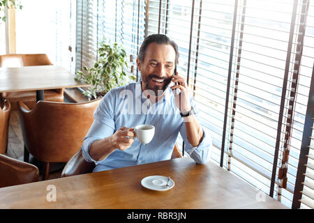 Reifer mann Händler am Tisch im Cafe sitzen tagsüber Holding Tasse trinken heißen Kaffee im Gespräch mit Freund auf dem Smartphone Lachen heiter Stockfoto