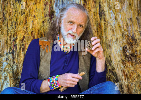 Gut aussehender älterer Mann in Hippie style gekleidet, in Rest lehnte sich gegen die sehr alte, beschädigte Pinie, holding Zweig und Pine Cone Stockfoto