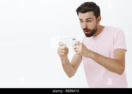 Nach bärtigen Mann spielt amüsantes Spiel über Smartphone, Handy mit beiden Händen konzentriert und an Handy Bildschirm mit coole App über grauer Hintergrund unterhalten aufgeregt Stockfoto