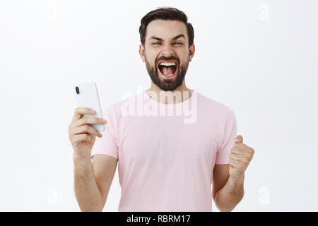 Aufgeregt und Triumphierte nach bärtigen Mann schreien Ja ballte die Fäuste in Sieg und Erfolg als Gewinnen in Telefon Spiel holding Smartphone feiern Sieg über den grauen Hintergrund zufrieden Stockfoto