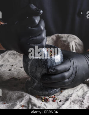 Koch in schwarzem Latex Handschuhe hält einen Stein Mörser mit einer Mischung aus Paprika, Kochen und Mahlen von Gewürzen Stockfoto