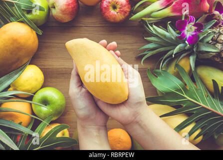 Close up weibliche Hand, die eine gelbe Mango auf die Gruppe der Früchte auf Holz Tisch Hintergrund Stockfoto
