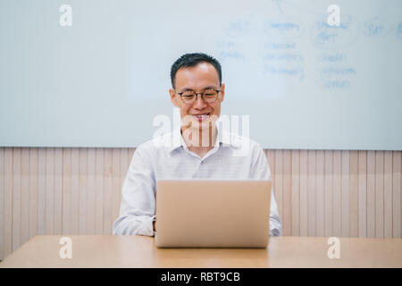 Eine junge chinesische Asian Business Mann arbeitet auf seinem Laptop in einem Konferenzraum in seinem Büro. Er lächelt, als er sich von seiner Arbeit sieht. Stockfoto