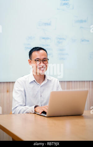 Eine junge chinesische Asian Business Mann arbeitet auf seinem Laptop in einem Konferenzraum in seinem Büro. Er lächelt, als er sich von seiner Arbeit sieht. Stockfoto