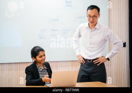 Eine vielfältige Business Team haben eine Besprechung Diskussion um einen Laptop im Büro. Ein chinesischer Mann und indische Frau talk intensiv miteinander. Stockfoto