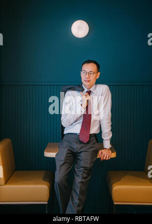Porträt eines asiatischen Geschäftsmann oder Professional (Rechtsanwalt, Bankier, etc) lehnte sich gegen einen Tisch in seinem Büro. Er ist groß, schlank und selbstbewusst. Stockfoto