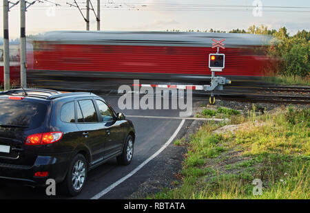 Beschleunigung Bewegungsunschärfe Red Train, der durch ein Bahnübergang mit Toren. Schwarzes Auto stehend vor dem bahnschranken auf eine asphaltierte Straße. Stockfoto