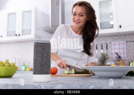 Glückliche Frau im Gespräch mit Voice Assistant beim Schneiden Gurken mit Messer Stockfoto